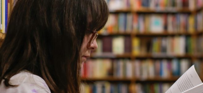 Dark haired woman reading a book, sitting leaning against stacks in a library.